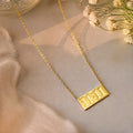 A gold chain necklace with a rectangular pendant engraved with '11:11', styled on a soft beige fabric with dried white flowers in the corner.