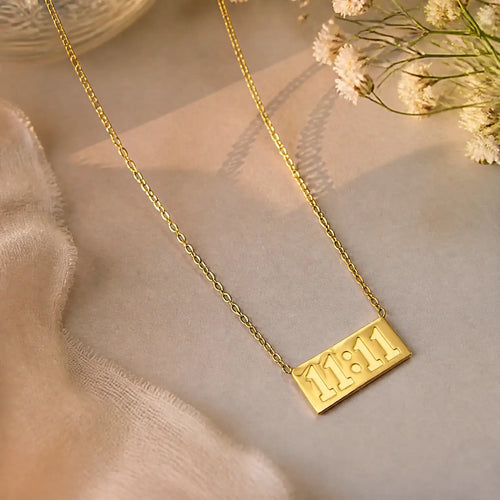 A gold chain necklace with a rectangular pendant engraved with '11:11', styled on a soft beige fabric with dried white flowers in the corner.
