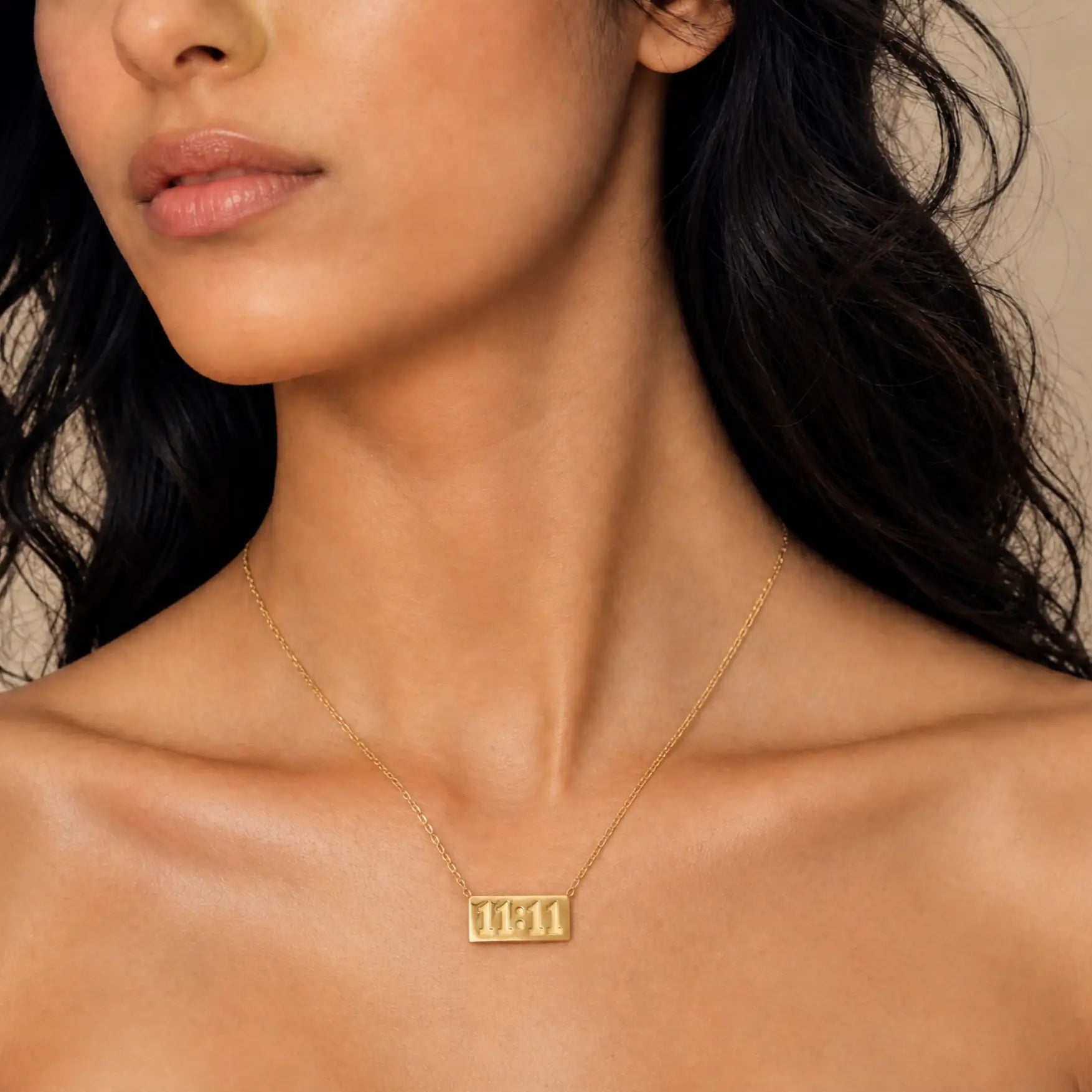 A close-up of a model wearing a delicate gold chain necklace with a rectangular pendant engraved with the Hebrew letters 'שם' (Shem), set against a warm beige background.