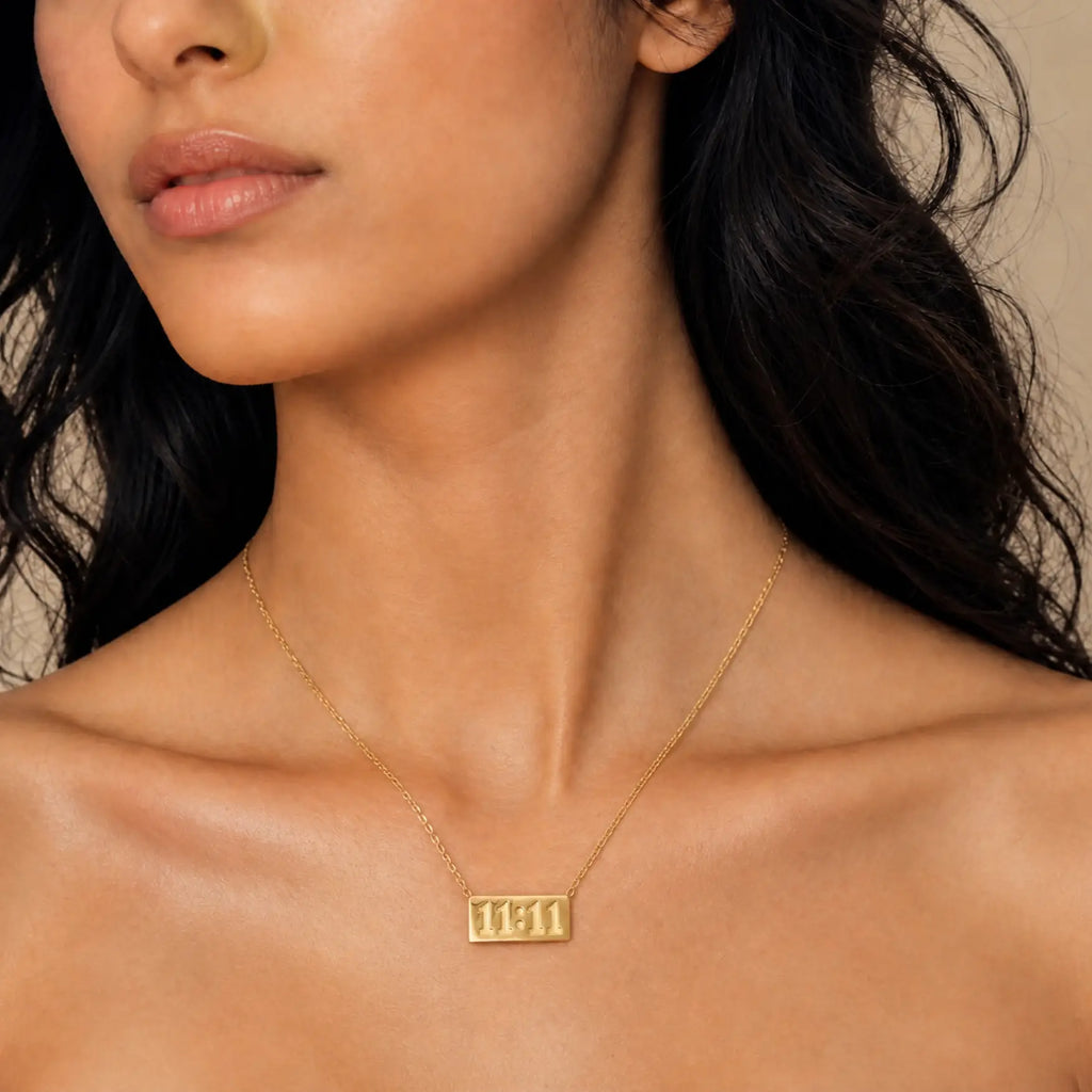 A close-up of a model wearing a delicate gold chain necklace with a rectangular pendant engraved with the Hebrew letters 'שם' (Shem), set against a warm beige background.