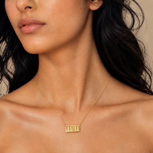 A close-up of a model wearing a delicate gold chain necklace with a rectangular pendant engraved with the Hebrew letters 'שם' (Shem), set against a warm beige background.