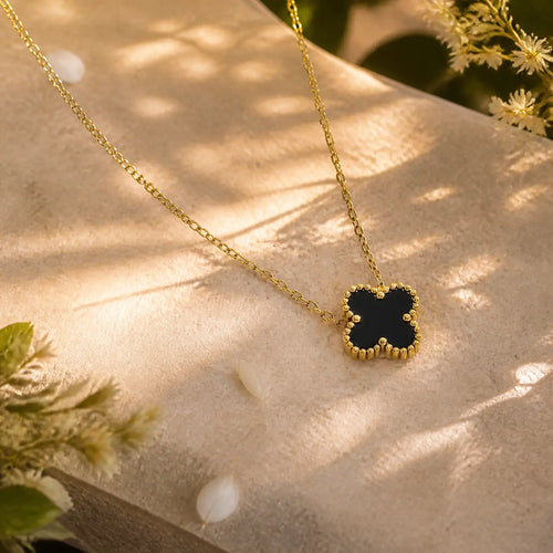 A gold chain necklace with a black clover-shaped pendant, styled on a textured beige stone surface with soft natural shadows and dried flowers.