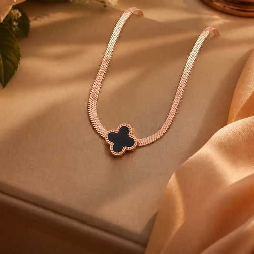 A delicate rose gold chain necklace with a clover-shaped pendant featuring a black center and a border of small clear stones, displayed on a beige satin fabric background.