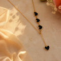 A gold-tone Y-necklace featuring four dangling heart charms with black inlay, styled on a beige textured surface with soft cream fabric and small flowers.
