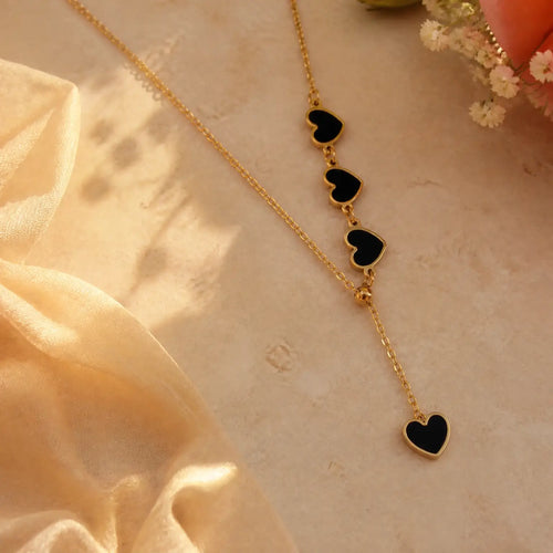 A gold-tone Y-necklace featuring four dangling heart charms with black inlay, styled on a beige textured surface with soft cream fabric and small flowers.