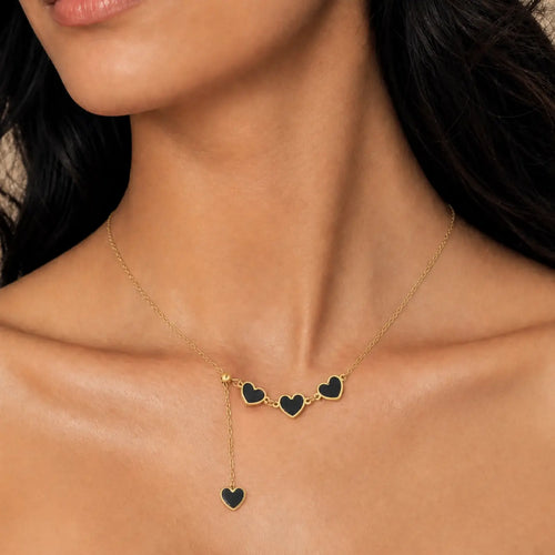 A close-up of a model wearing a delicate gold necklace with three small heart charms and a dangling heart pendant, each filled with black enamel.