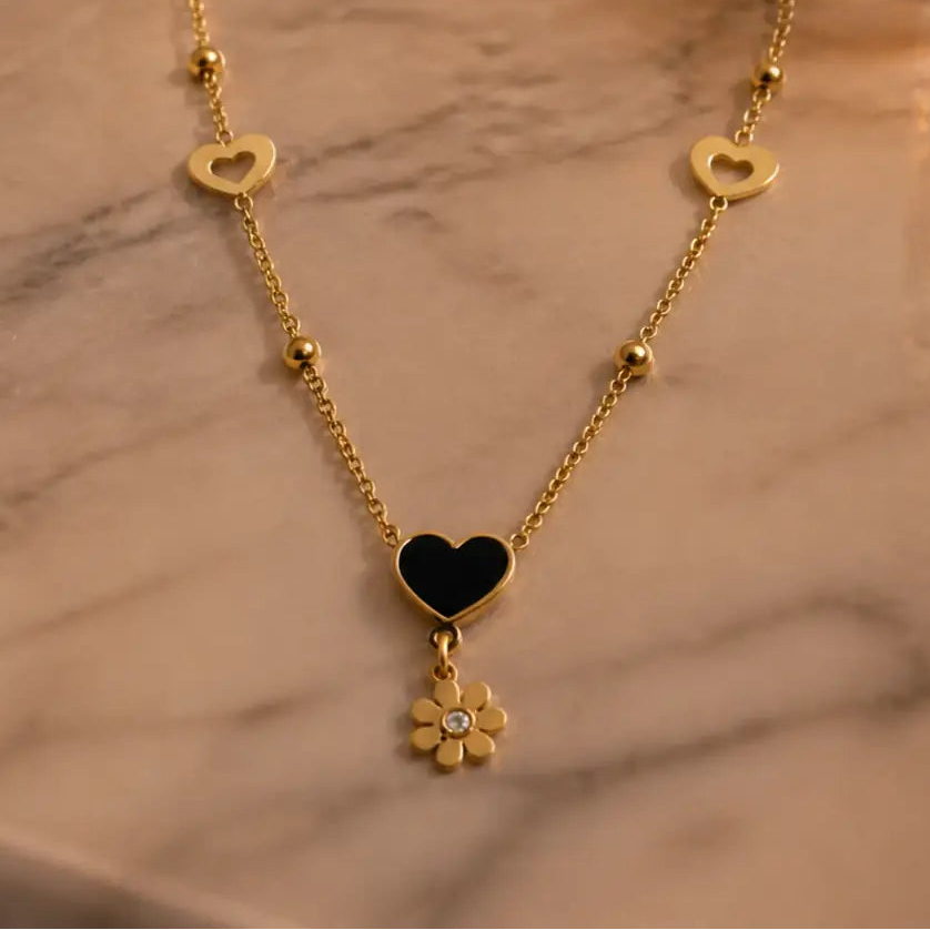 A gold chain necklace with a central black heart pendant and a dangling flower charm, displayed on a light marble surface.