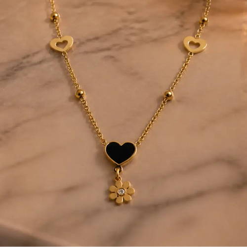A gold chain necklace with a central black heart pendant and a dangling flower charm, displayed on a light marble surface.