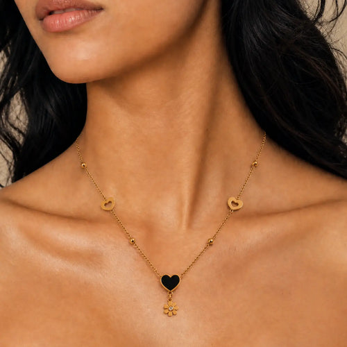 A close-up of a model wearing a delicate gold chain necklace with a central black heart pendant, flanked by small gold heart and flower charms.
