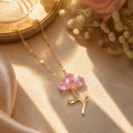 A gold-tone floral pendant necklace with pink gemstone petals and small green leaf accents, styled on a beige satin surface with dried flowers and warm sunlight.