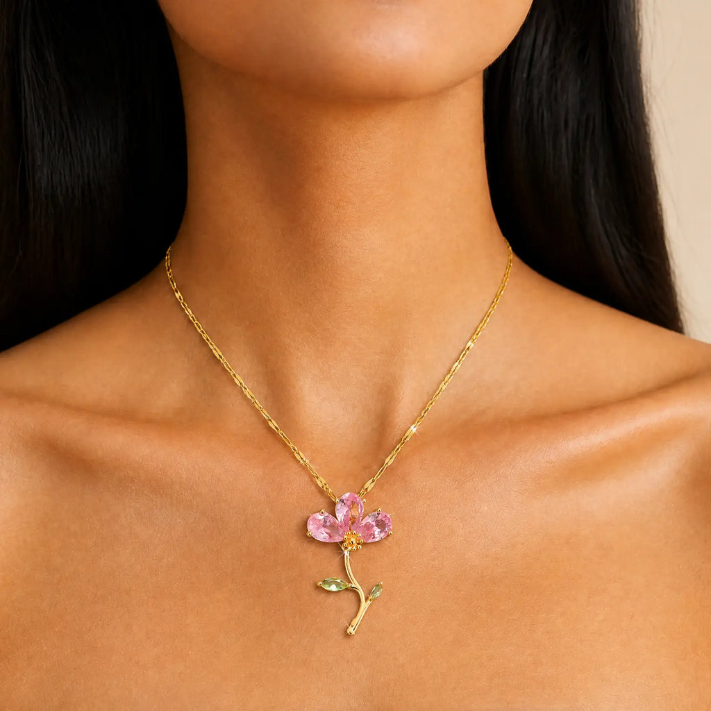 A close-up of a model's neck and shoulders wearing a delicate gold chain necklace with a flower pendant featuring pink petals and green leaves.