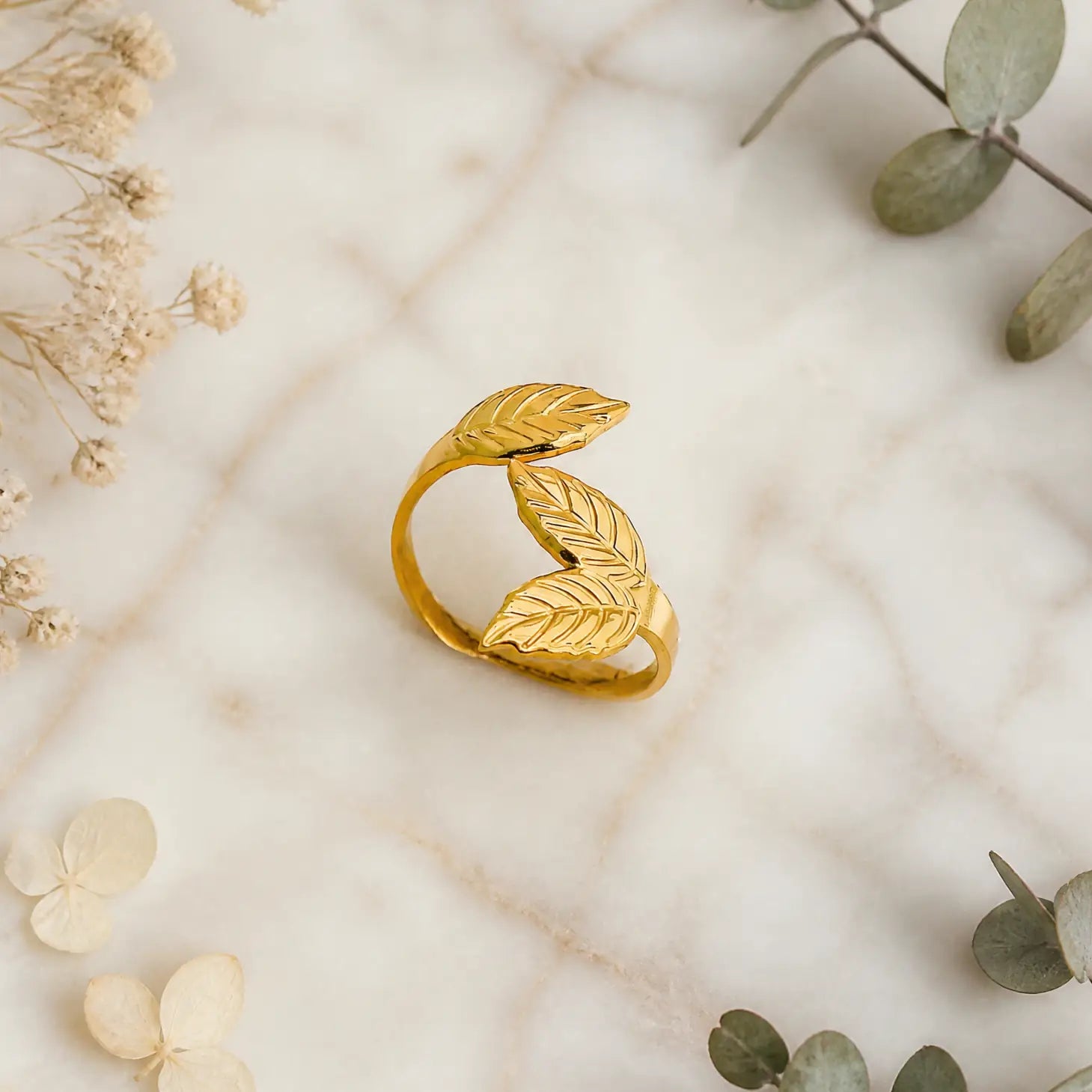 A gold leaf-shaped ring with two overlapping leaf designs, displayed on a light marble surface with dried flowers and green leaves as decorative props.