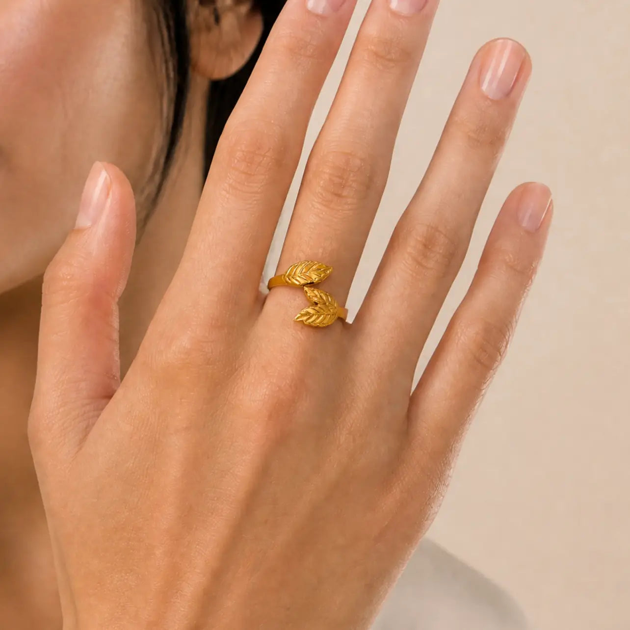 A close-up of a hand wearing a gold ring shaped like two overlapping leaves, shown against a soft beige background.