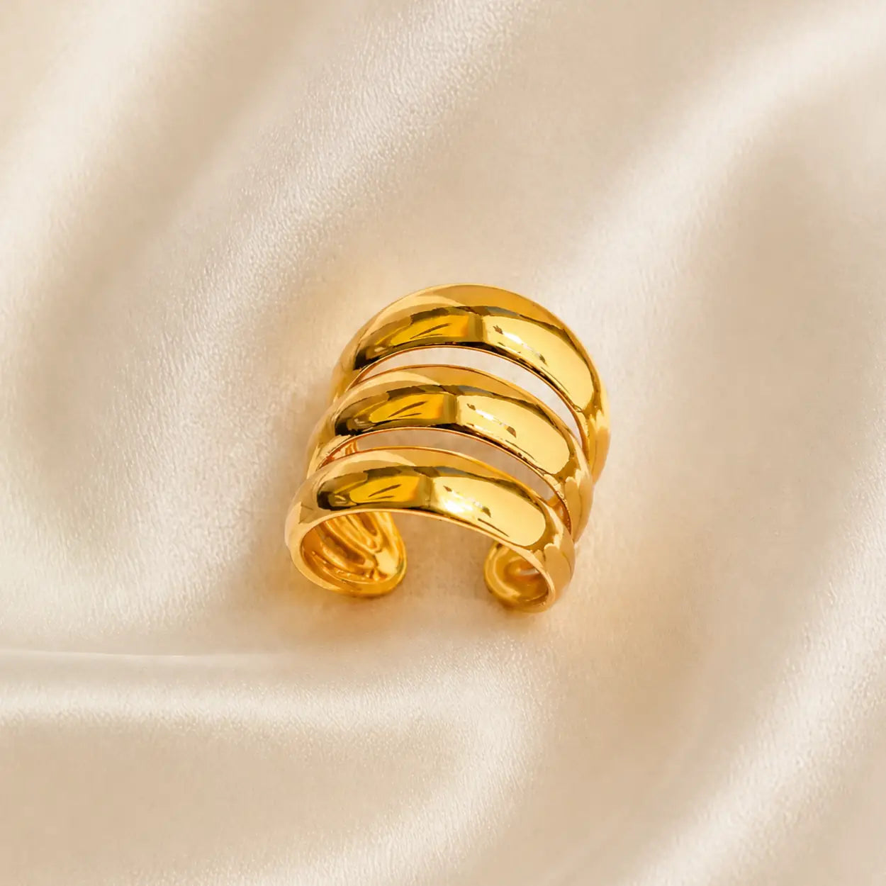 A polished gold wrap ring with three horizontal bands, displayed on a soft, cream-colored satin fabric background.