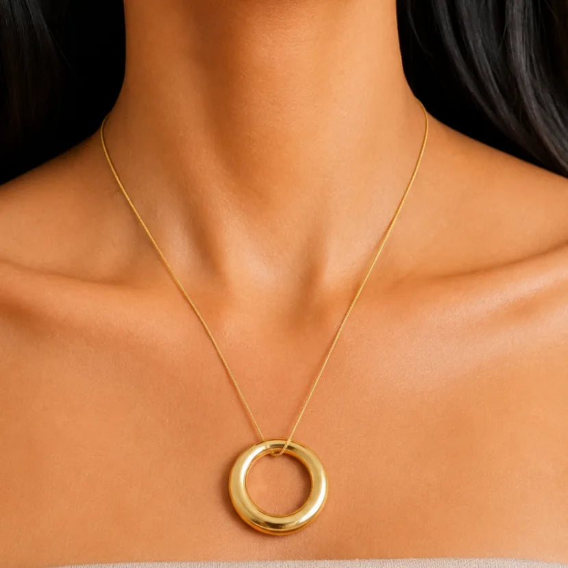 A close-up of a model's neck and shoulders wearing a delicate gold chain necklace with a thick, polished gold ring pendant.