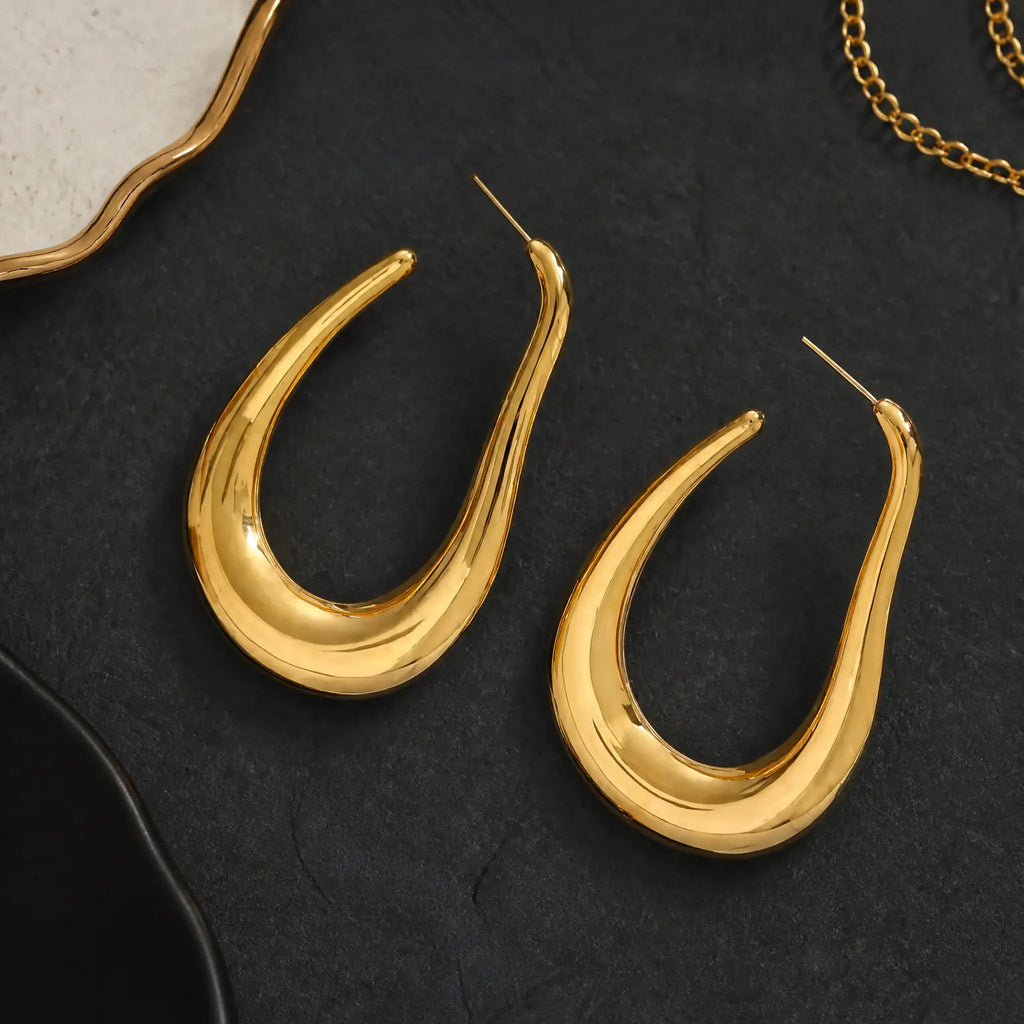 A pair of gold teardrop-shaped hoop earrings with a polished finish, displayed on a dark textured surface with decorative jewelry props.