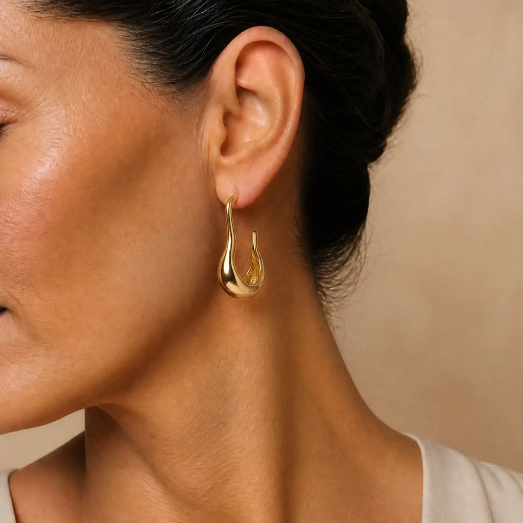 A close-up of a woman's ear wearing a polished gold teardrop-shaped hoop earring against a warm beige background.
