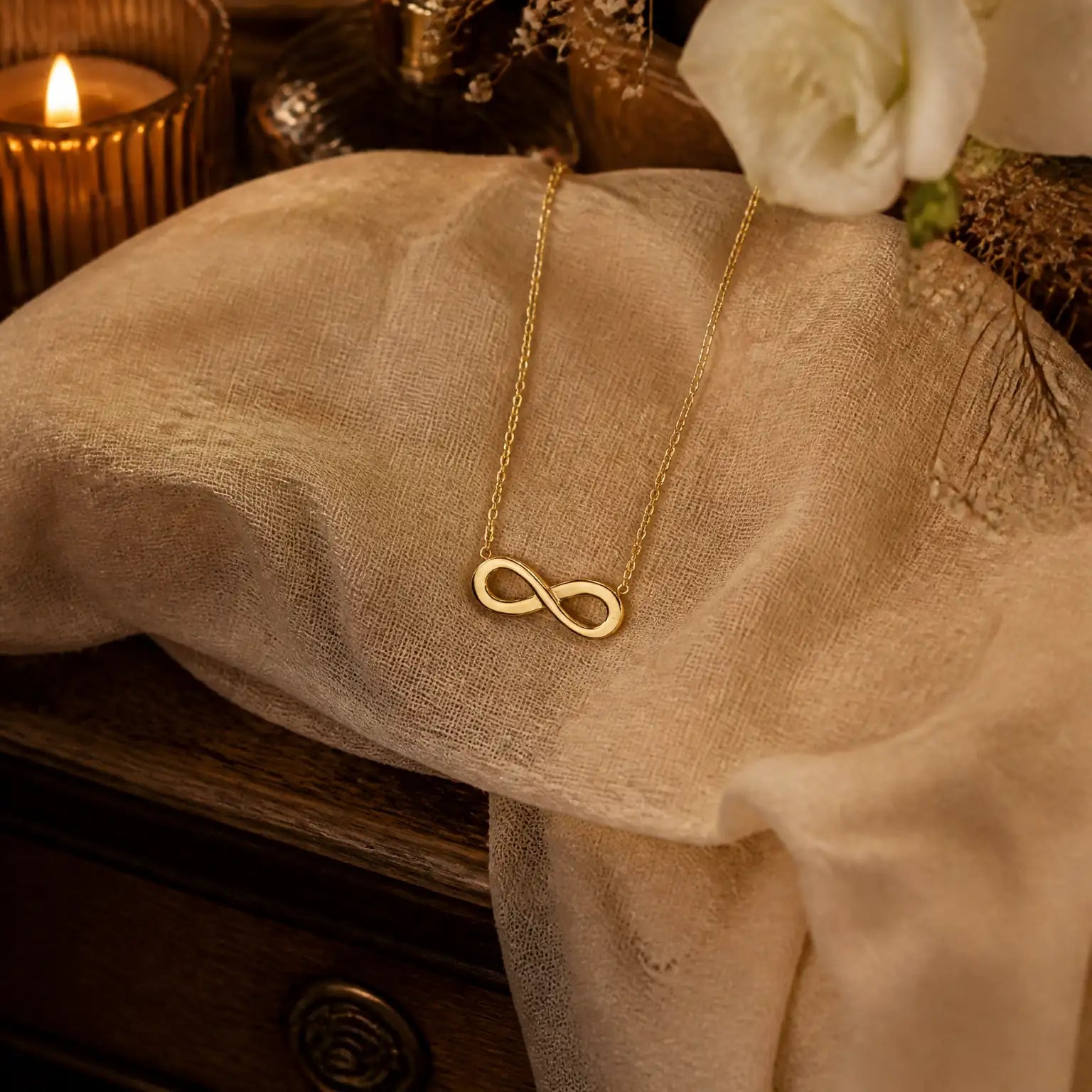 A delicate gold infinity necklace draped over a beige fabric surface, styled with a lit candle, a white rose, and a dark wooden surface in a warm, cozy setting.
