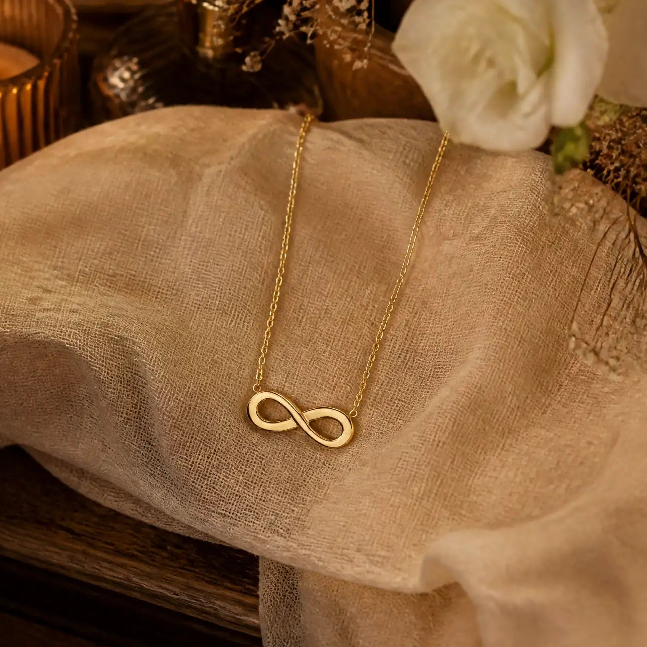 A delicate gold infinity necklace draped over a beige fabric surface, styled with a lit candle, a white rose, and a dark wooden surface in a warm, cozy setting.