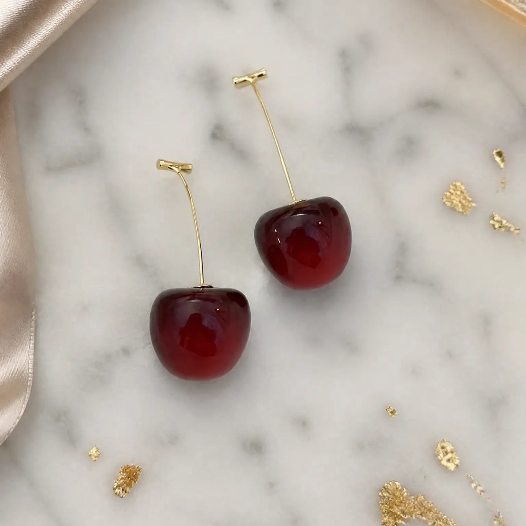 A pair of cherry-shaped drop earrings with glossy deep red bodies and gold-tone stems, displayed on a white marble surface with gold leaf and satin ribbon accents.
