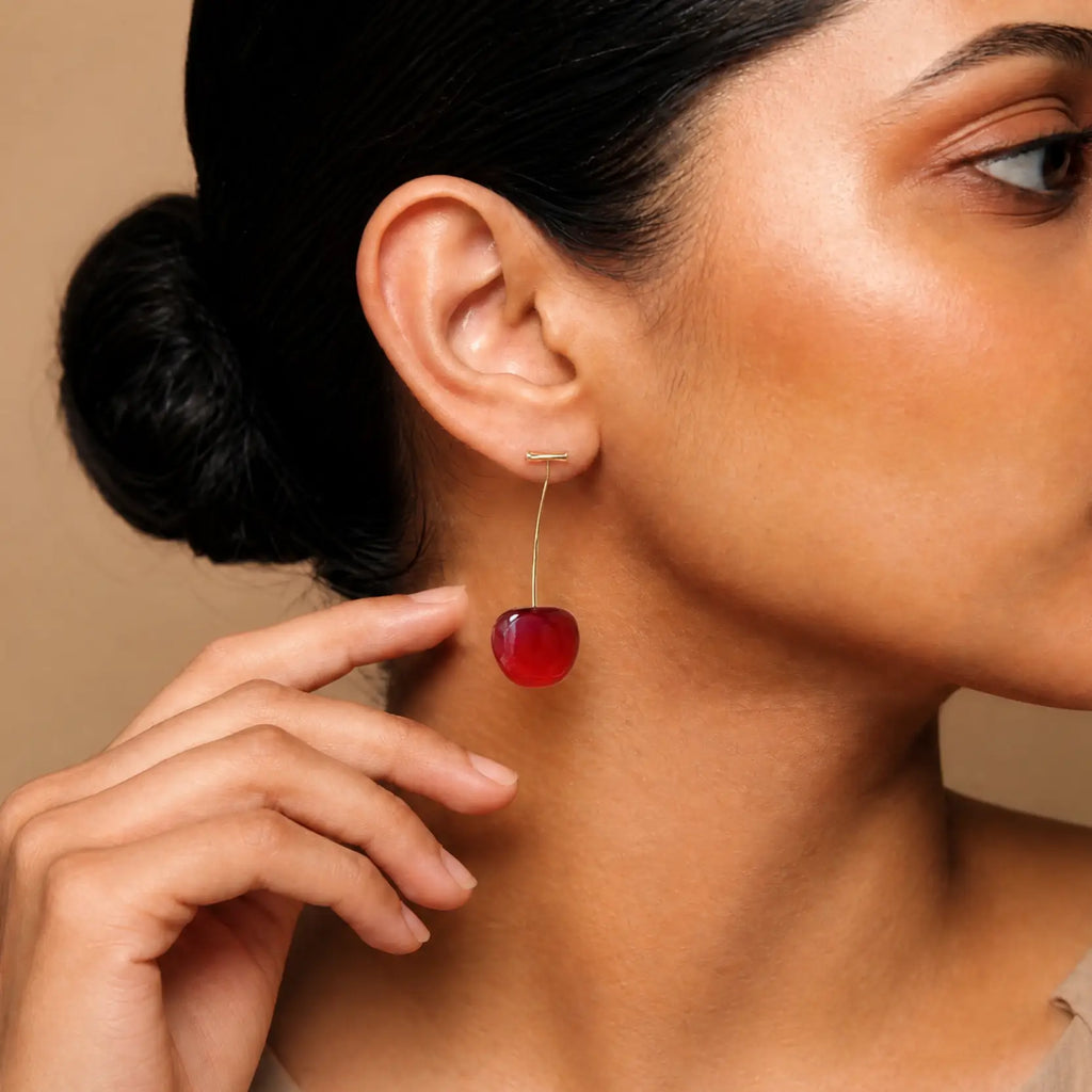 A close-up of a woman wearing a dangling cherry earring with a polished red cherry charm suspended from a thin gold-tone wire, shown against a warm beige background.
