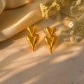 A pair of gold heart-shaped earrings displayed on a beige fabric surface with soft cream-colored dried flowers and draped satin in the background.