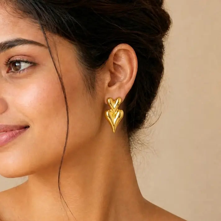 A close-up of a woman's ear wearing a shiny gold heart-shaped dangle earring against a soft beige background.