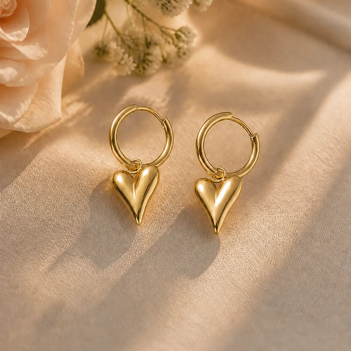 A pair of gold hoop earrings with dangling heart charms, displayed on a beige fabric surface with soft shadows and a pale rose in the background.