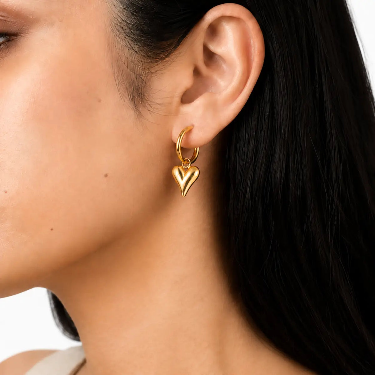 A close-up of a model's ear wearing a small gold hoop earring with a dangling polished gold heart charm.