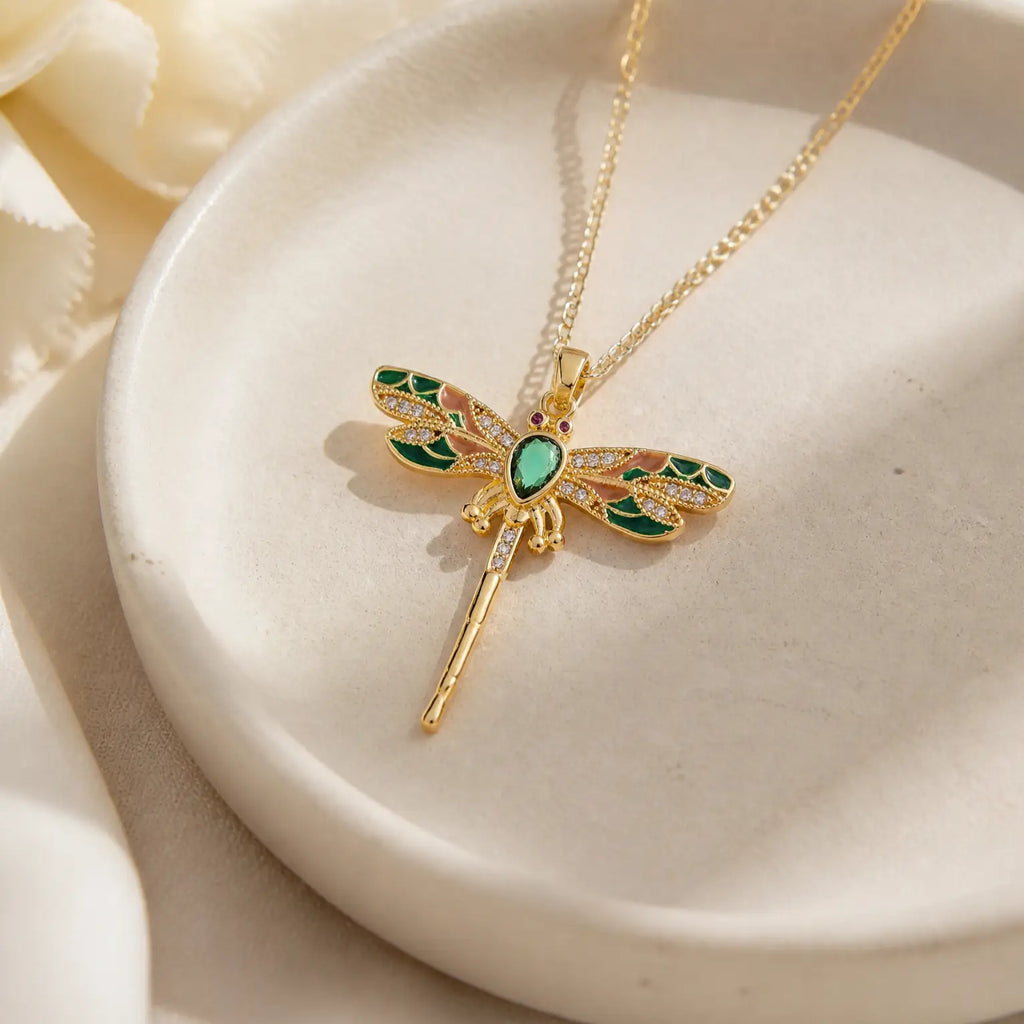 Gold dragonfly pendant necklace with green and gold enamel wings, displayed on a white ceramic dish.