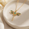 Gold dragonfly pendant necklace with green and gold enamel wings, displayed on a white ceramic dish.