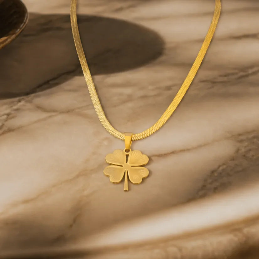 A gold necklace with a four-leaf clover pendant, displayed on a beige marble surface with soft shadows.