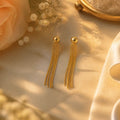A pair of gold tassel drop earrings with spherical studs, displayed on a warm beige marble surface with soft floral and fabric props.