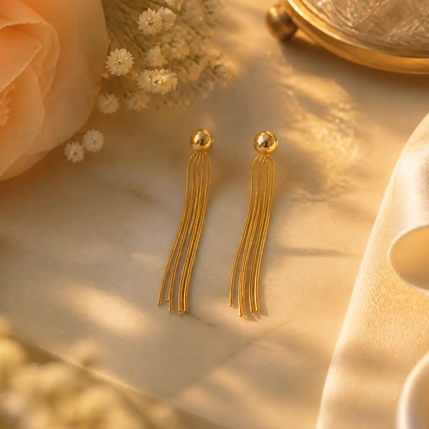 A pair of gold tassel drop earrings with spherical studs, displayed on a warm beige marble surface with soft floral and fabric props.