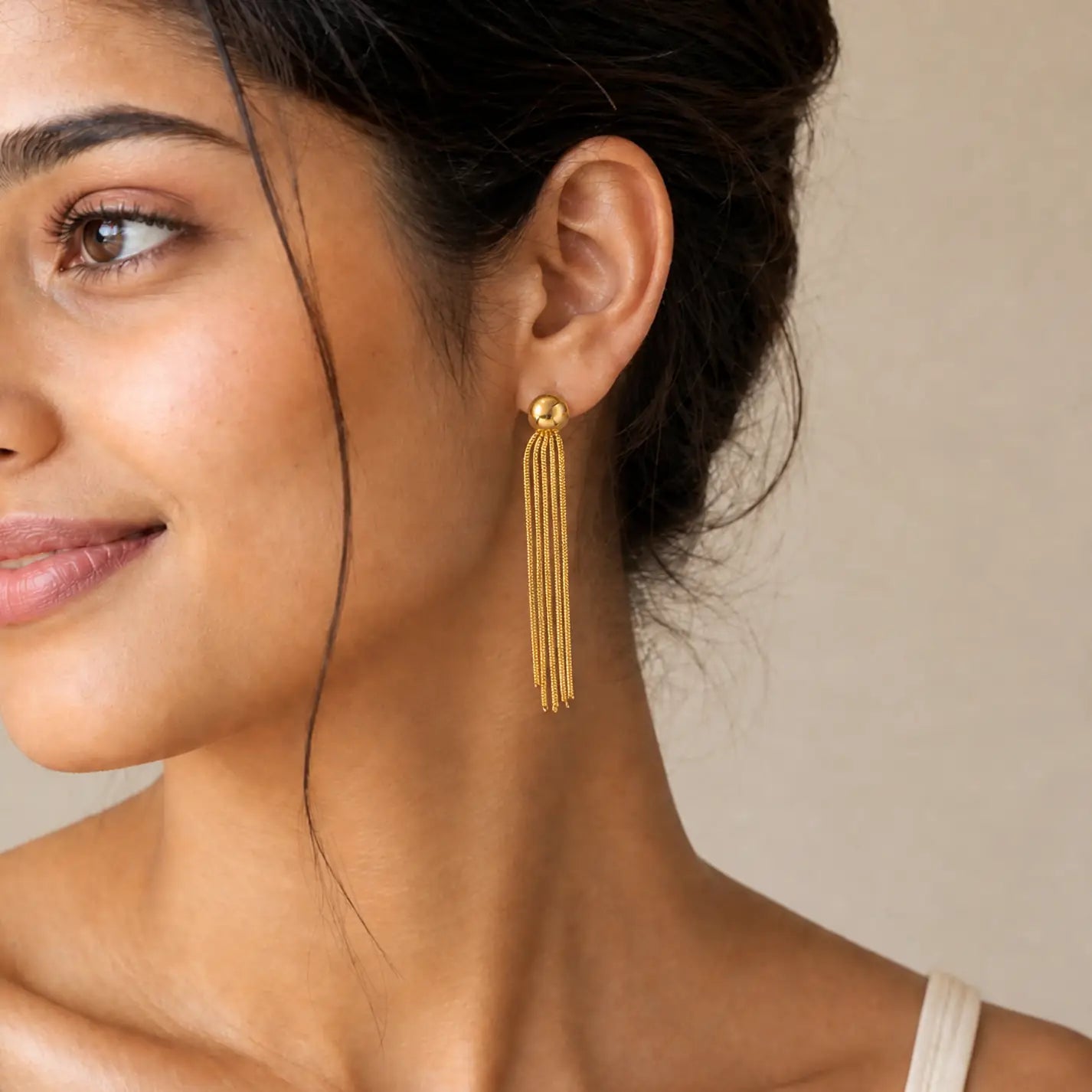 A close-up of a woman wearing a single gold dangle earring with multiple thin vertical strands hanging from a round stud, against a soft beige background.