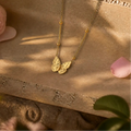 A delicate gold necklace with a butterfly pendant lies on a textured stone surface, surrounded by soft pink flower petals and green leaves in warm natural light.