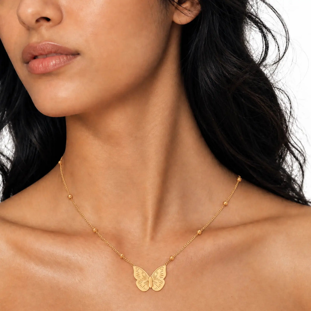 A close-up of a model wearing a delicate gold chain necklace with a small butterfly pendant at the center, set against a clean white background.