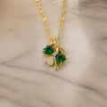 A gold-plated shamrock pendant necklace with two green heart-shaped emerald-cut stones and one clear crystal-cut stone, resting on a light beige marble surface.