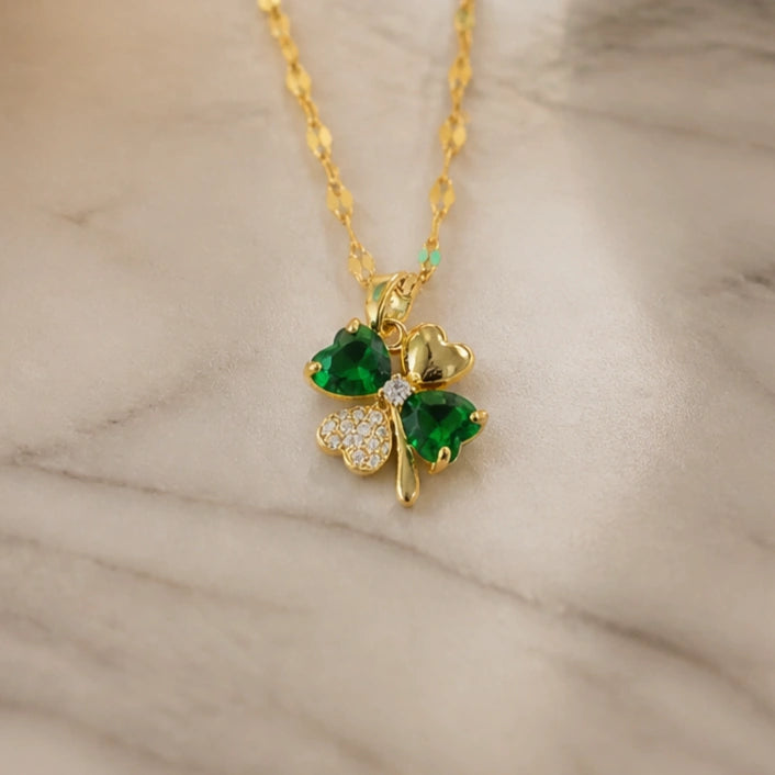 A gold-plated shamrock pendant necklace with two green heart-shaped emerald-cut stones and one clear crystal-cut stone, resting on a light beige marble surface.