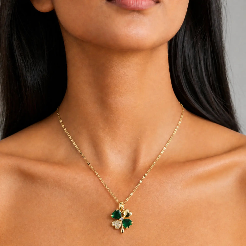 A close-up of a model wearing a delicate gold chain necklace with a four-leaf clover pendant. The pendant features two green emerald-cut stones and one clear round-cut stone, set against a neutral gray background.