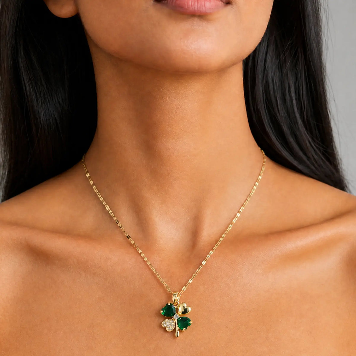 A close-up of a model wearing a delicate gold chain necklace with a four-leaf clover pendant. The pendant features two green emerald-cut stones and one clear round-cut stone, set against a neutral gray background.