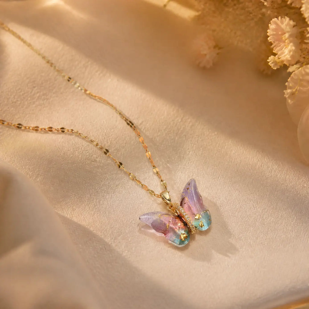 A delicate gold chain necklace with a butterfly pendant featuring iridescent pastel wings in pink, purple, and blue, displayed on a soft beige fabric background with dried flowers.