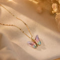 A delicate gold chain necklace with a butterfly pendant featuring iridescent pastel wings in pink, purple, and blue, displayed on a soft beige fabric background with dried flowers.