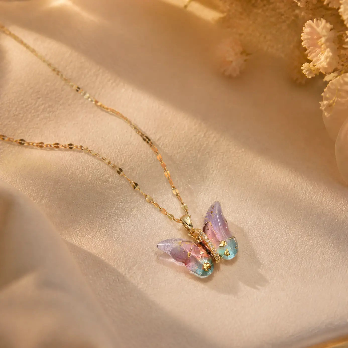 A delicate gold chain necklace with a butterfly pendant featuring iridescent pastel wings in pink, purple, and blue, displayed on a soft beige fabric background with dried flowers.