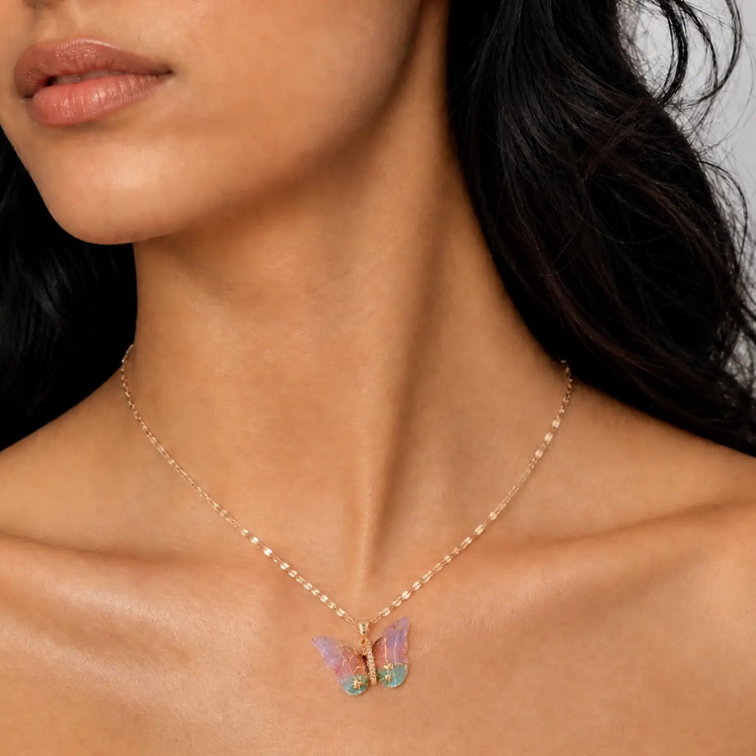 A close-up of a model wearing a delicate gold chain necklace with a butterfly pendant. The butterfly features iridescent pastel wings in pink, purple, and green, set against a soft neutral background.