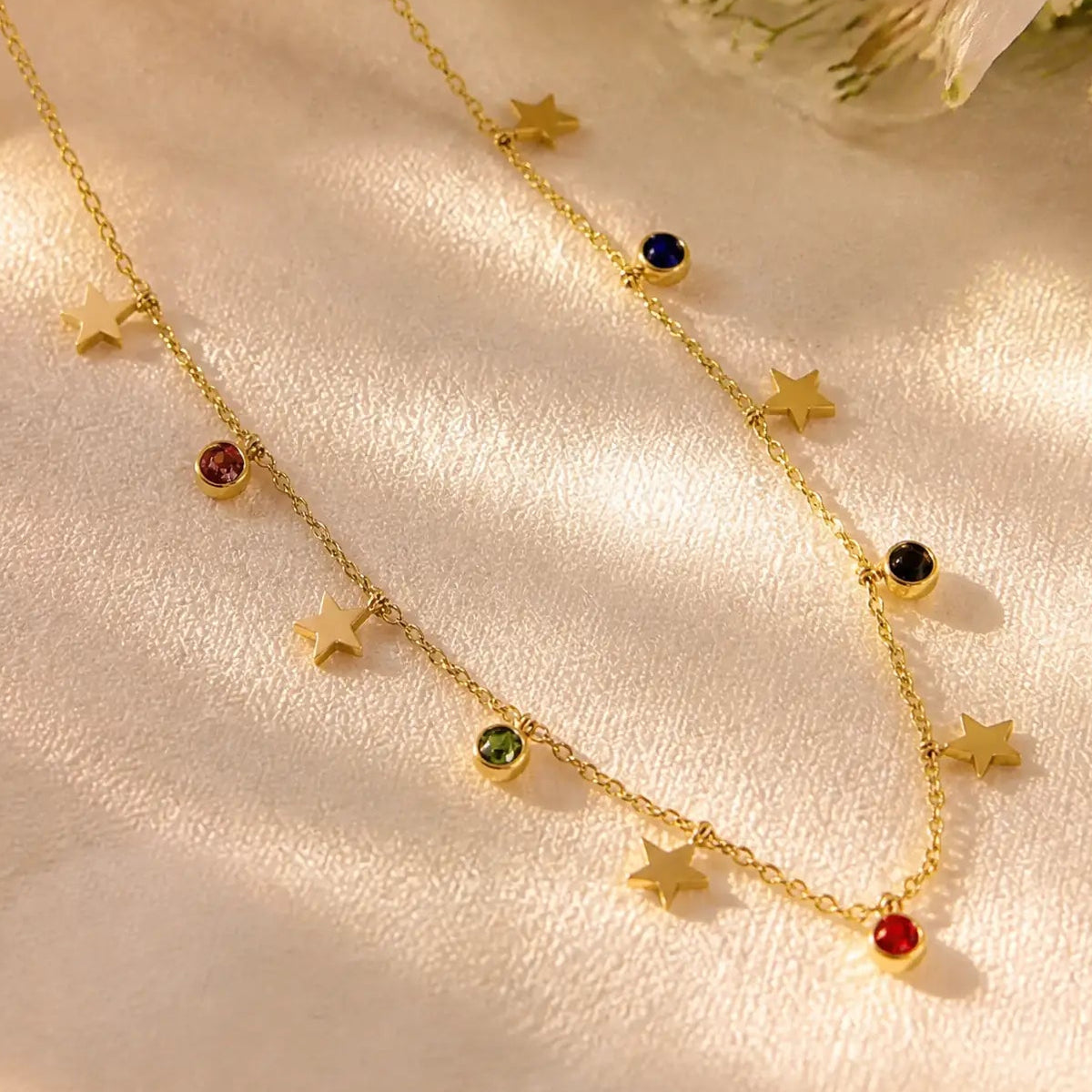 A delicate gold chain necklace with multiple dangling star charms and small round gemstone accents in red, green, blue, and black, displayed on a soft cream-colored fabric background with warm sunlight shadows.