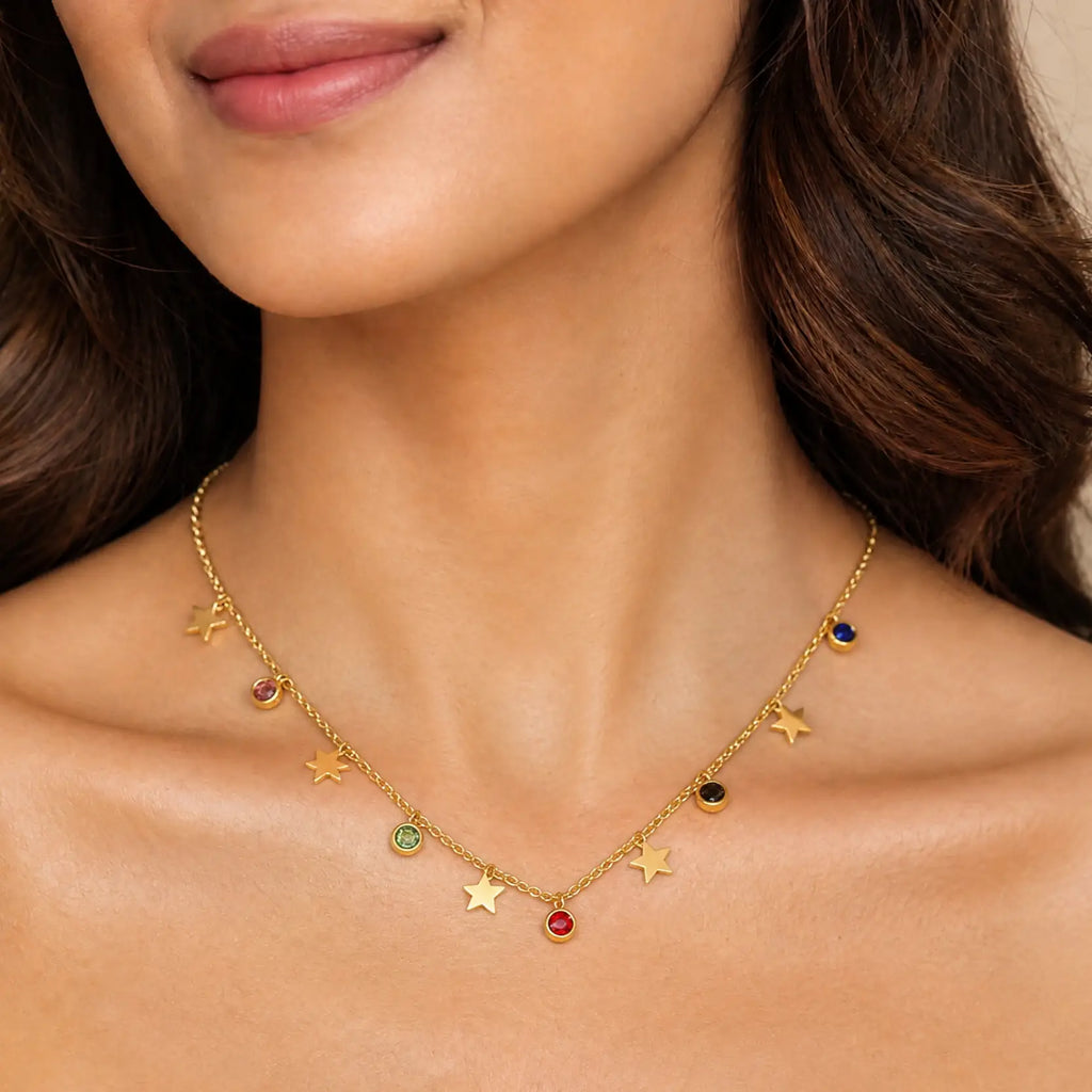 A close-up of a woman wearing a delicate gold necklace with star and moon charms, each accented with small colorful gemstones.