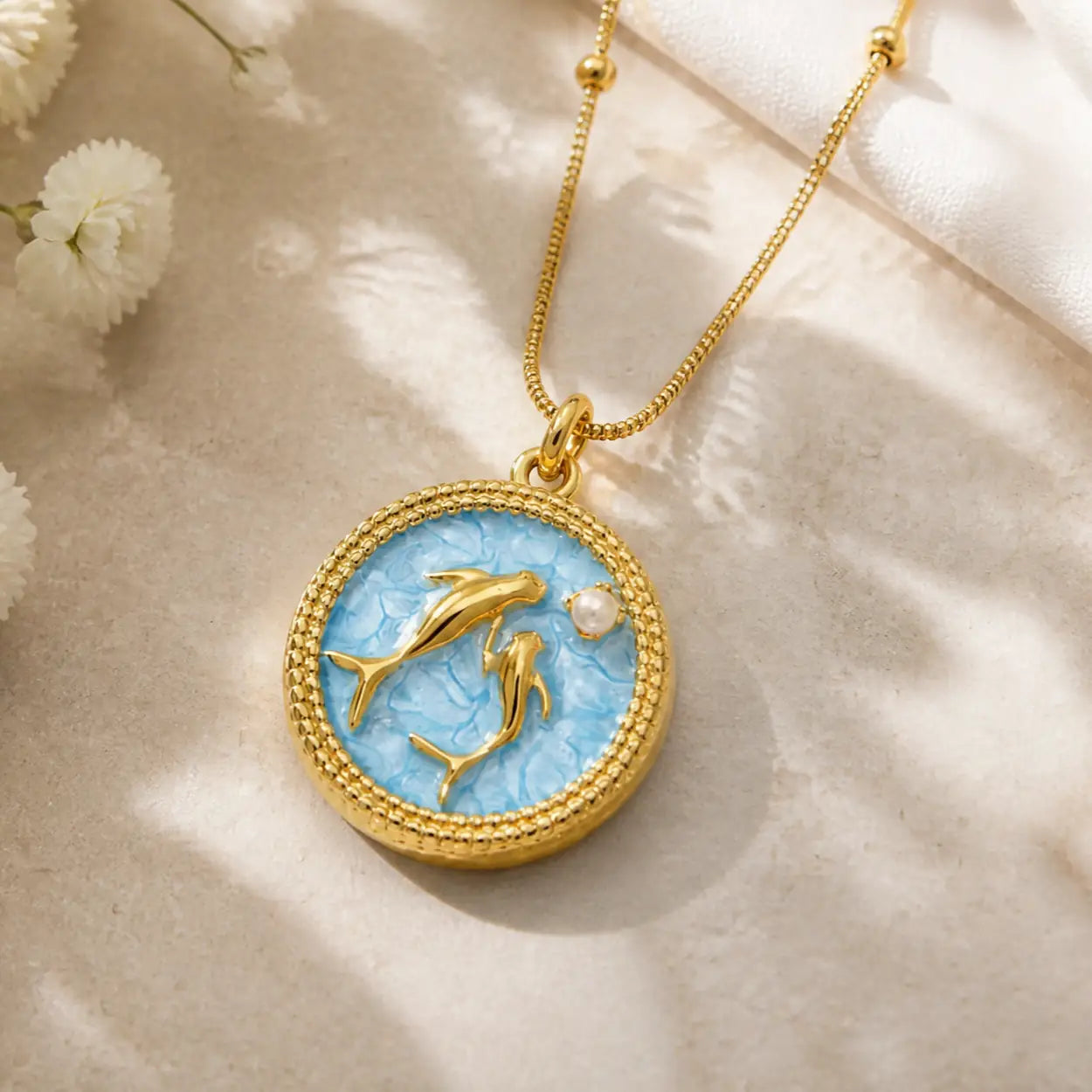 A gold pendant necklace featuring two gold dolphins on a textured light blue circular background, accented with a small white pearl, displayed on a beige fabric surface with soft shadows.
