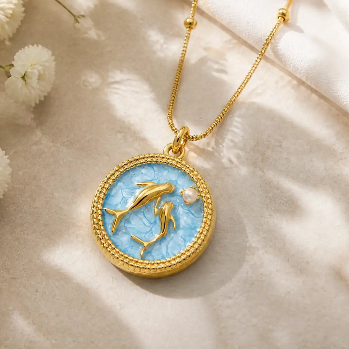 A gold pendant necklace featuring two gold dolphins on a textured light blue circular background, accented with a small white pearl, displayed on a beige fabric surface with soft shadows.