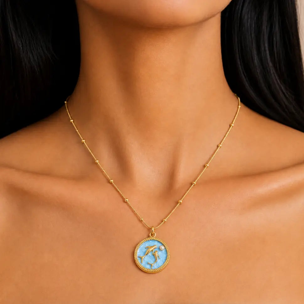 A gold necklace with a circular pendant featuring a light blue background and a gold dolphin design, shown worn on a model's neck.
